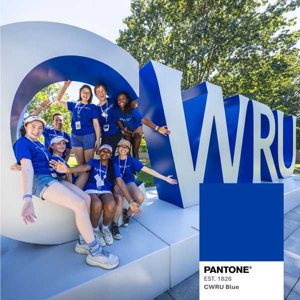 A group of students posing by a large gray and blue sign that spells, “CWRU” with a Pantone-style color swatch labeled “CWRU Blue.”