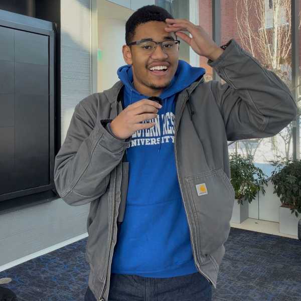 A student wearing a blue hoodie and glasses holding a small microphone in one hand, with his other hand on his forehead.