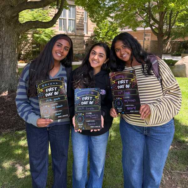 Three CWRU students pose on the lawn in front of Adelbert Hall holding filled-out “First Day of Class” signs.