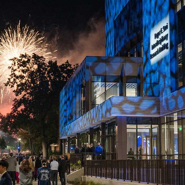 The Roger E. Susi Interdisciplinary Research Building illuminated in blue spotlights at night, with fireworks lighting up the sky behind it.
