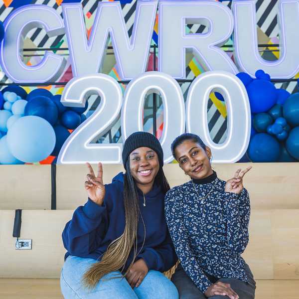 Two women posing with peace sign hand gestures in Tinkham Veale University Center in front of a balloon and letter display reading, “CWRU 200.”