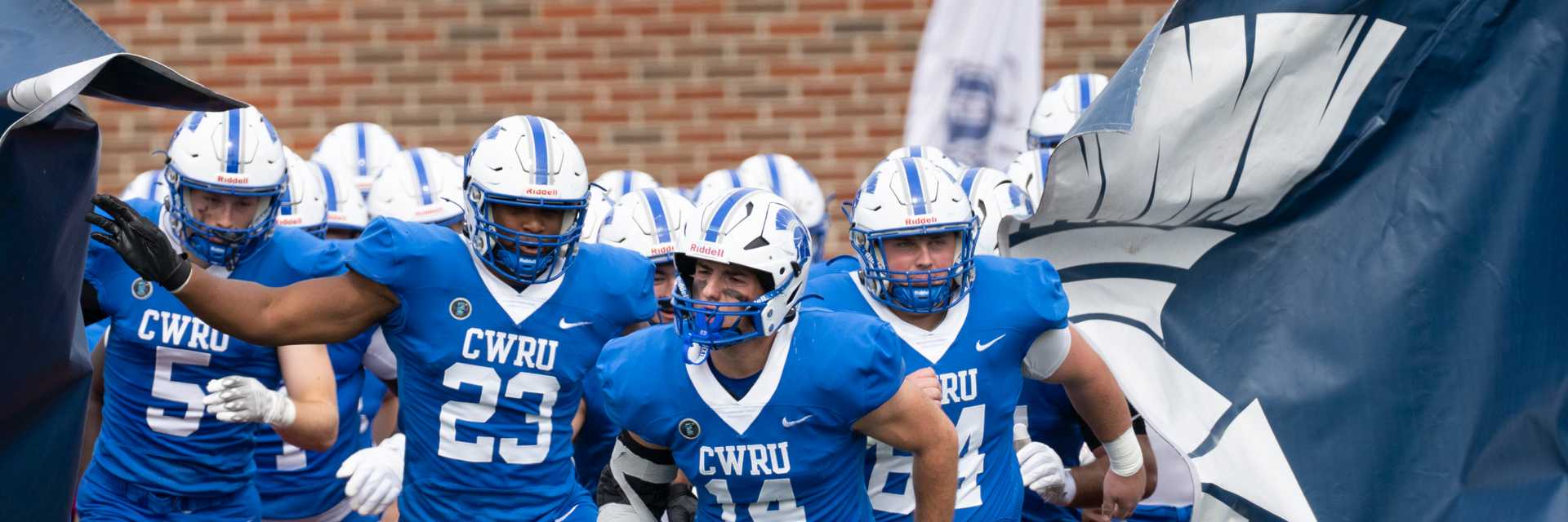 CWRU football players run onto the field through a Spartan banner